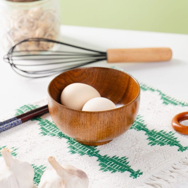 Zwei Eier in einer rustikalen Holzschüssel. Im Hintergrund sind ein Holzschneebesen und Essstäbchen zu sehen.