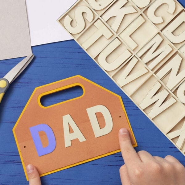 Kinderhände basteln ein Vatertagsschild aus großen Holzbuchstaben. Auf dem Schild steht „DAD“.