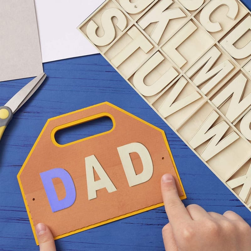 Kinderhände basteln ein Vatertagsschild aus großen Holzbuchstaben. Auf dem Schild steht „DAD“.