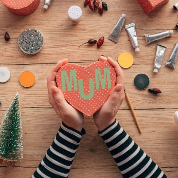 Hände halten eine herzförmige Schachtel mit der Aufschrift „MUM“. Bastelmaterial liegt auf einer Holzoberfläche und umgibt die Hände.