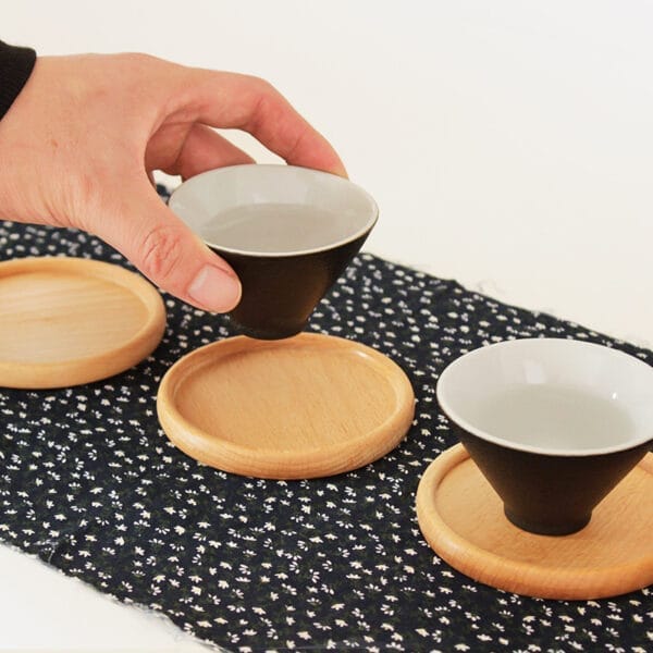 Hand stellt eine schwarz-weiße Teetasse auf einen runden Holzuntersetzer. Weitere Untersetzer und Tassen sind sichtbar.