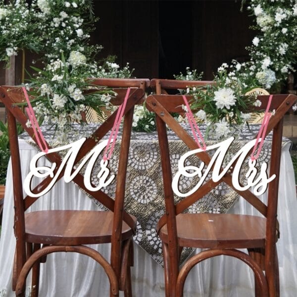 An Hochzeitsstühlen hängen hölzerne „Mr. & Mrs.“-Schilder, geschmückt mit weißen Blumen.