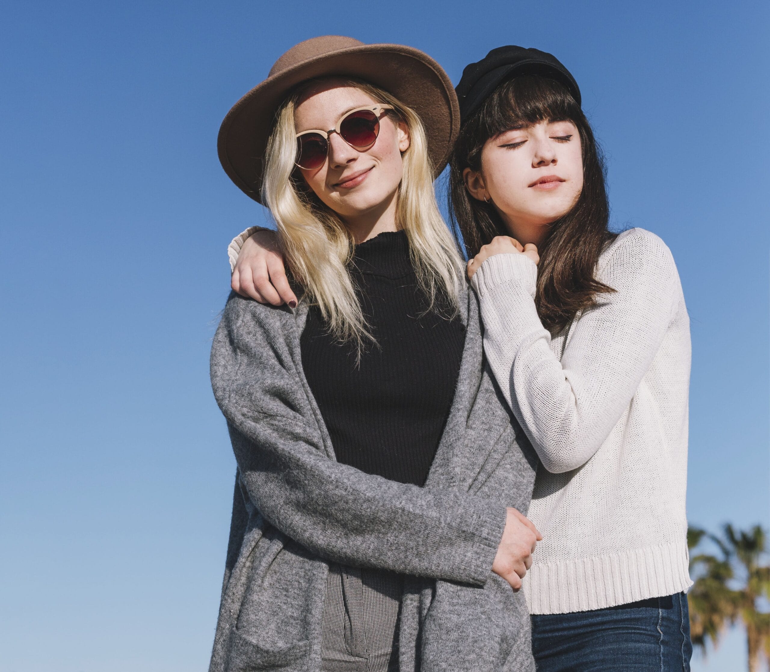 Zwei junge Freundinnen umarmen sich und tragen modische Hüte und Pullover vor einem strahlend blauen Himmel.