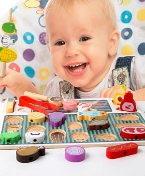 Fröhliches Kleinkind, das mit einem hölzernen Grillspielzeugset spielt. Großhandel Holzspielzeug.