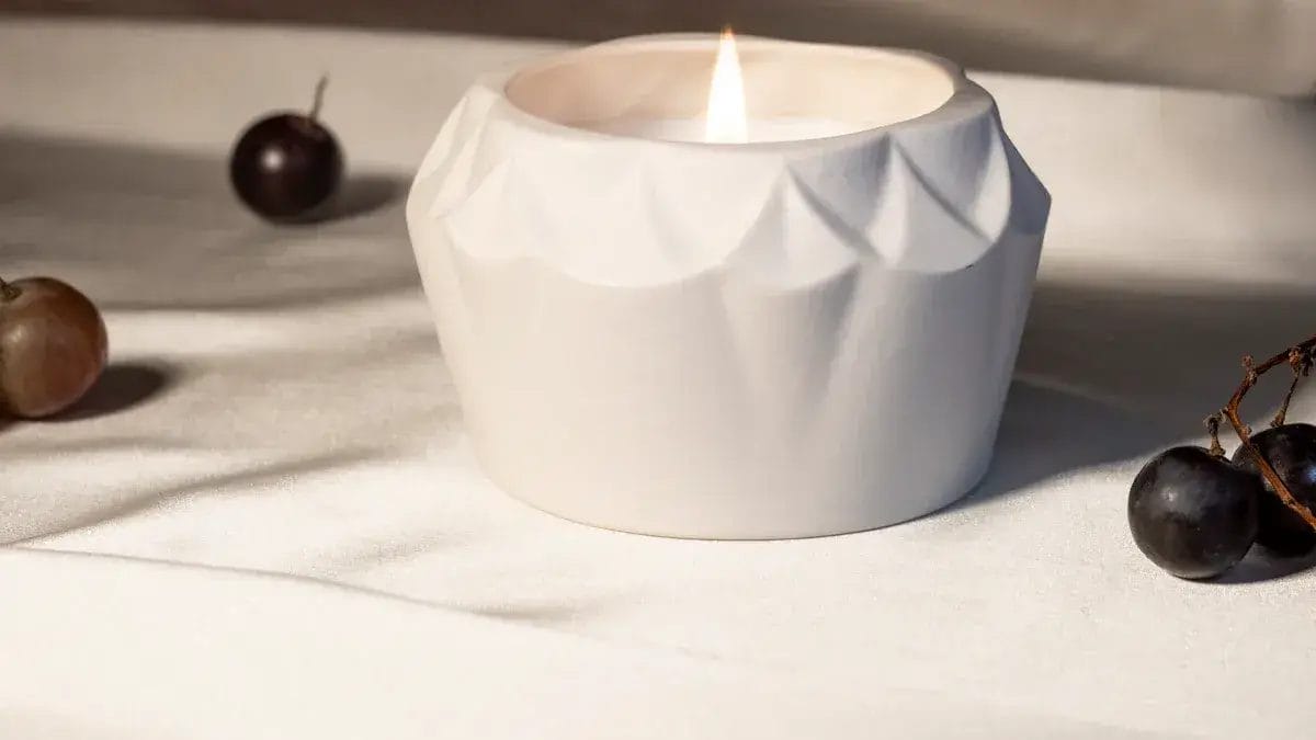 Weiße Kerze in einem dekorativen Kerzenhalter mit Trauben auf einem weißen Tuch.