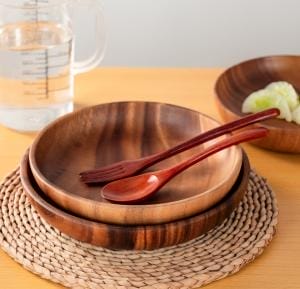 Flache Holzschale mit Salat, Gabel und Löffel auf geflochtenem Untersetzer. Nachhaltige Küchenschüssel.