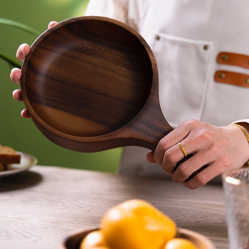 Holzschale mit Griff, gehalten von einer Person über einem Tisch mit Früchten.